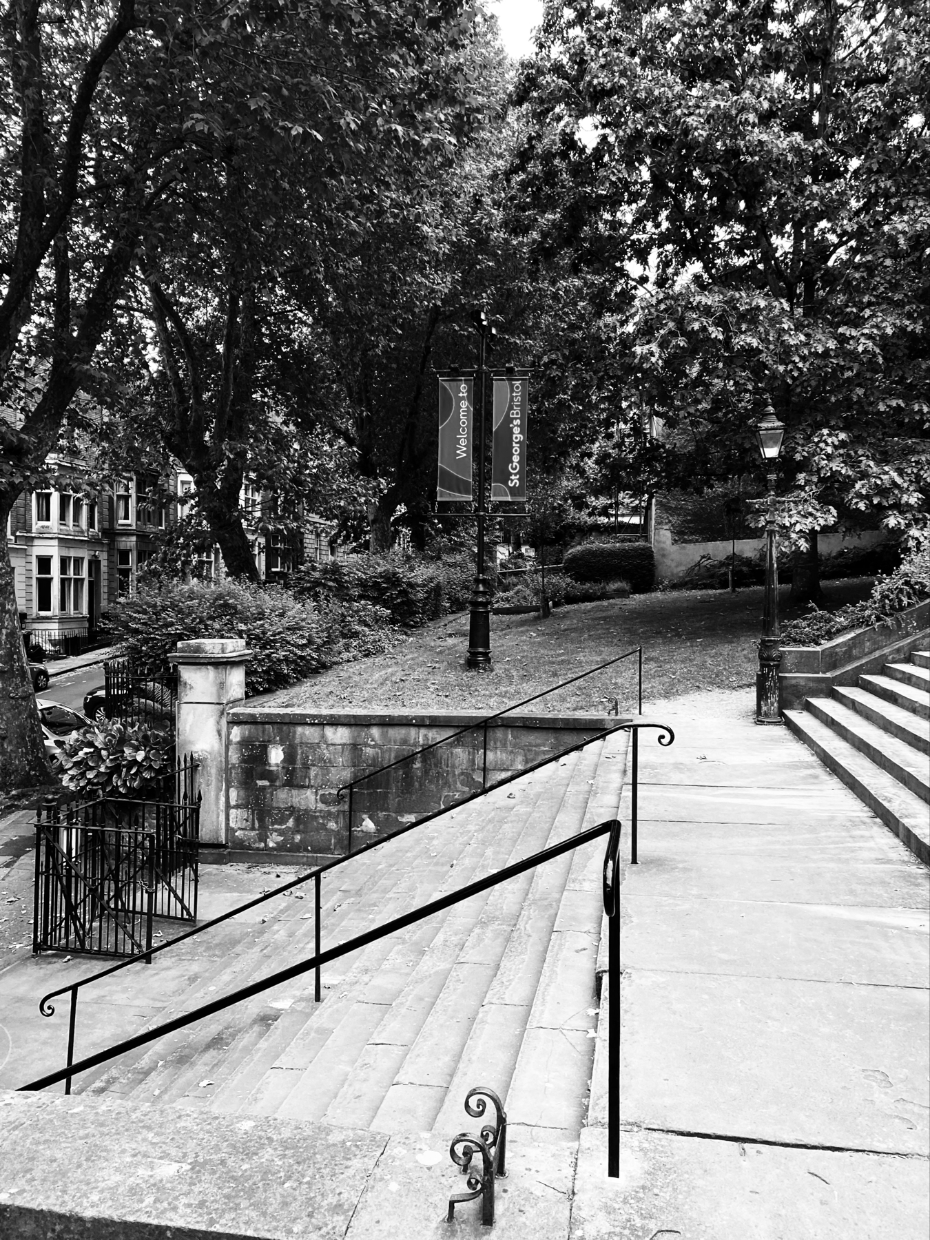 Black and white photo of a stone stairway and railings, surrounded by trees and historic buildings. A sign reads Welcome to St George's Bristol. The scene also includes iron railings and a lamp post.