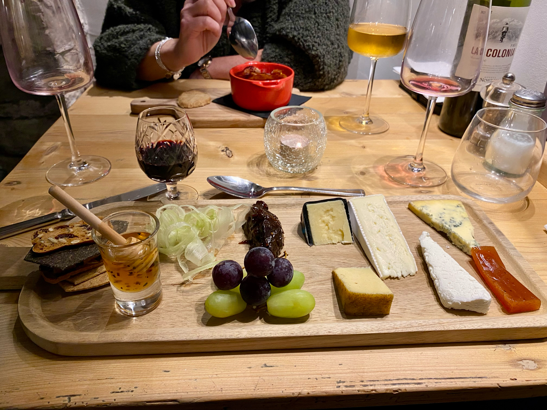 A cheese platter with various cheese types, grapes, crackers, honey, and pickled items, accompanied by glasses of port and dessert wine. A person is dining in the background.