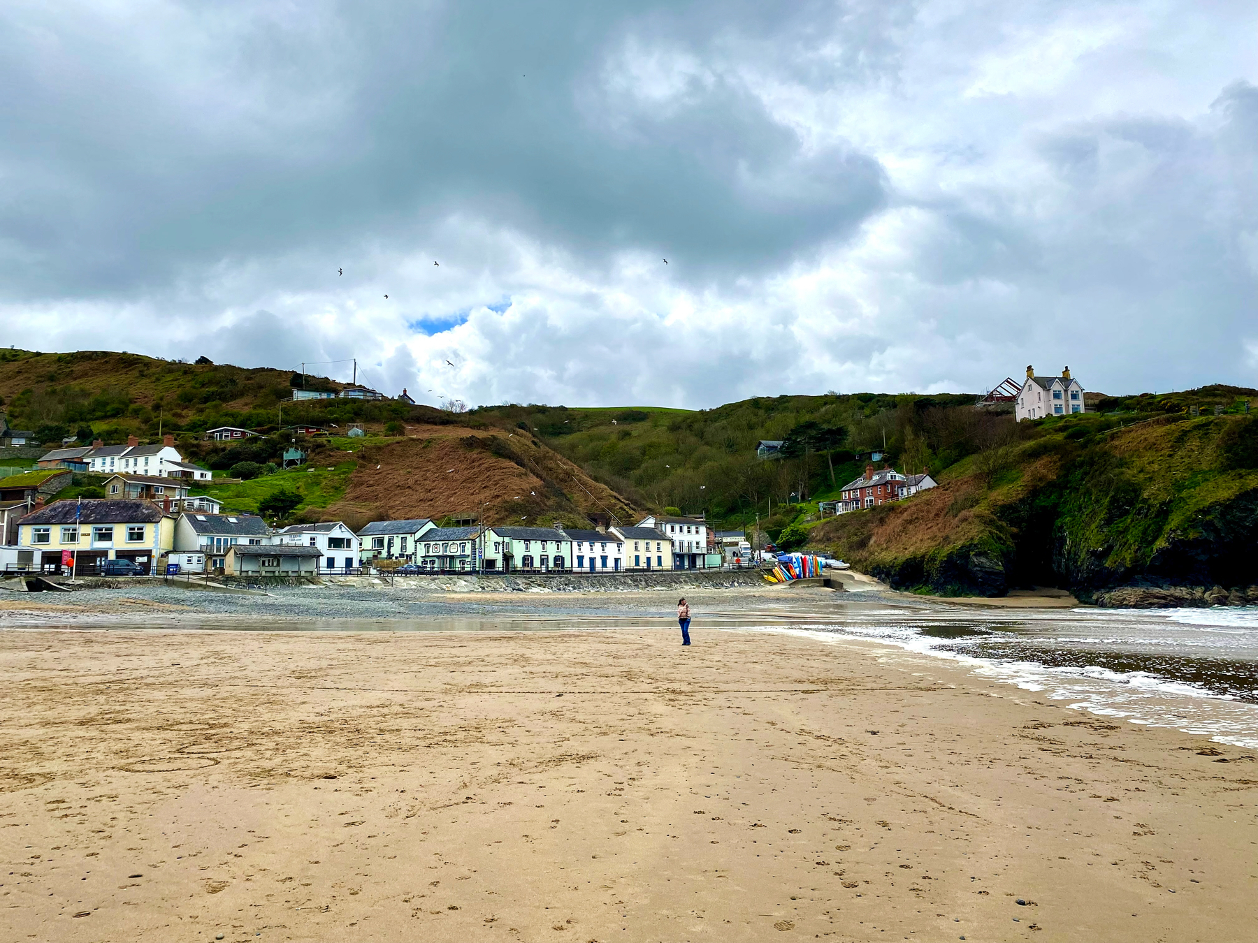 Sandy beach with a lone figure walking, coastal buildings nestled on the hillside, dark clouds overhead.