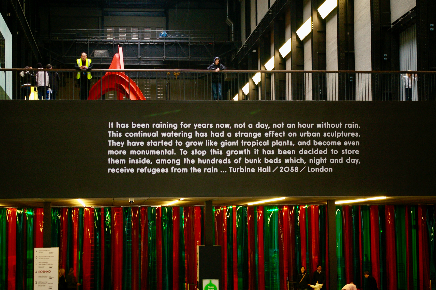 Interior view of a building with an artistic installation: a text panel with a narrative about continuous rain and its effects on urban sculptures, set in the future, above a colorful fringe curtain. People are visible on an upper balcony.