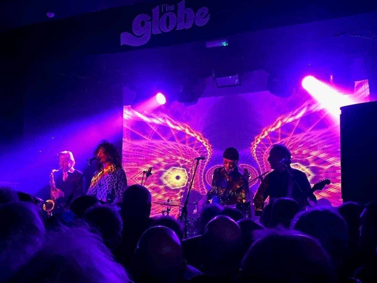 A live music performance at The Globe with musicians on stage, including a saxophonist and guitarists, amid colorful stage lighting and a crowd of spectators in the foreground
