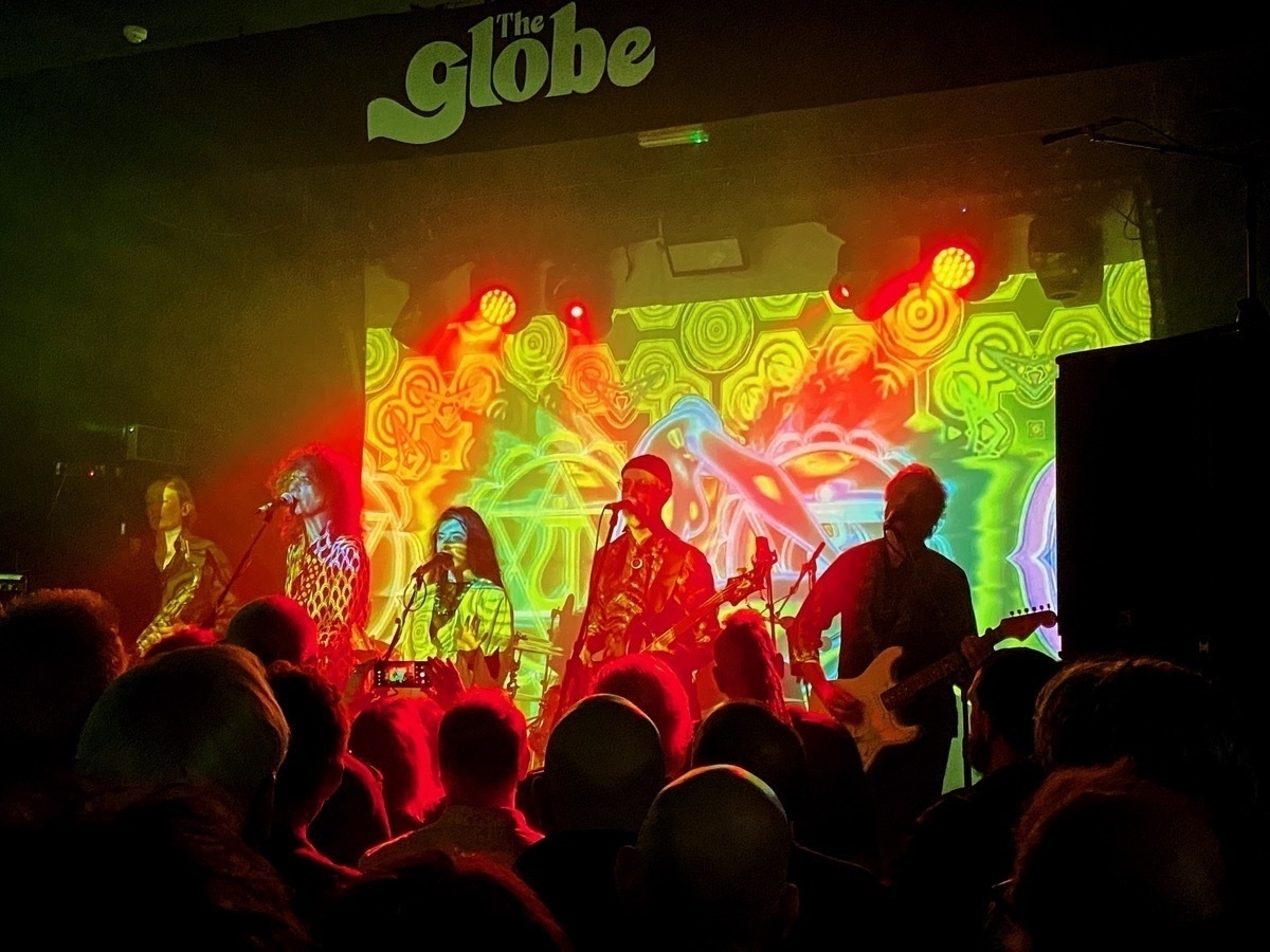 A live band performing on stage at The Globe with vibrant psychedelic background projections and an audience in the foreground