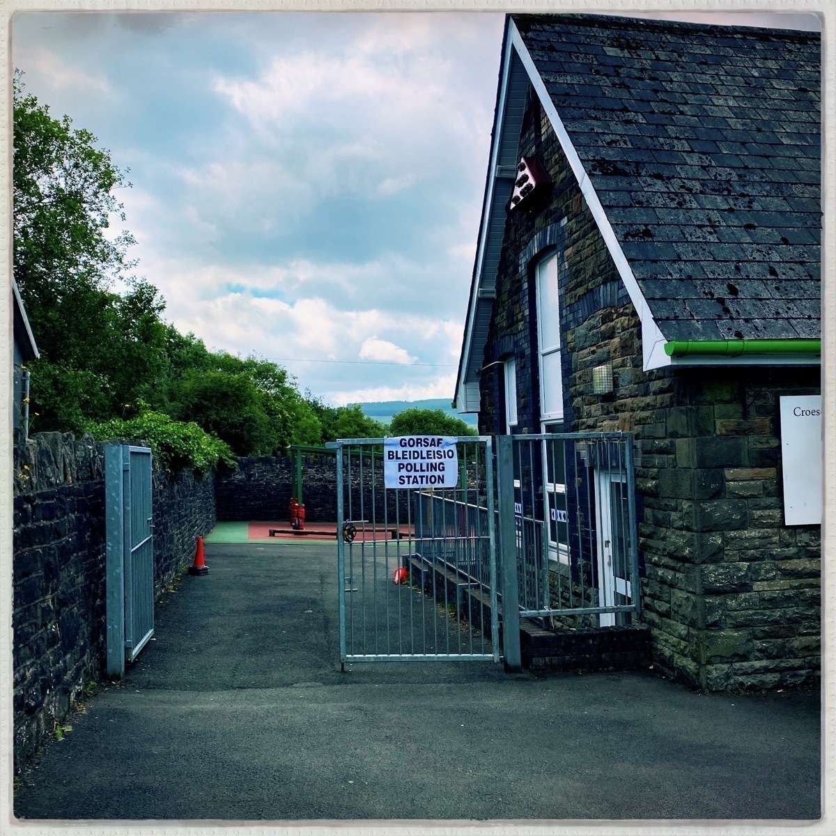 A stone building with a slate roof, serving as a polling station, is situated behind a gated entrance with surrounding greenery under a cloudy sky