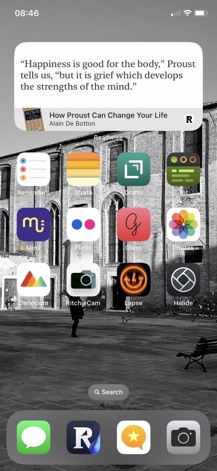Screenshot of an iPhone screen displaying a variety of apps, a widget with a literary quote from Marcel Proust, and a black and white background image of a public square with a person walking and birds on the ground.
