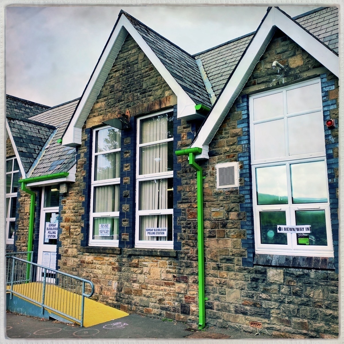 A stone building with large windows and green gutters appears to be an entrance to a school, as indicated by the signs on the doors and windows.