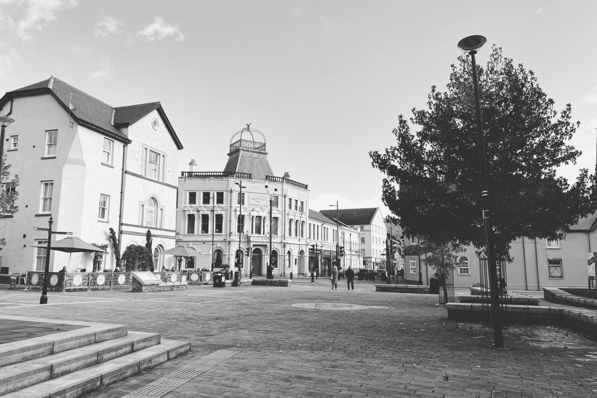 Black and white photo of a town square with historic buildings, a few pedestrians, lamp posts, and a tree.