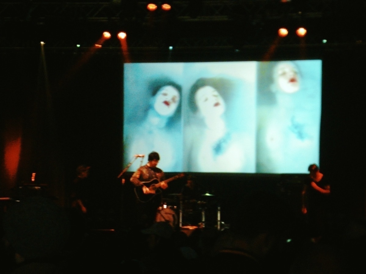 Darkly lit band on stage with backdrop of bright screens with hazy pictures of a person wearing bright red lipstick