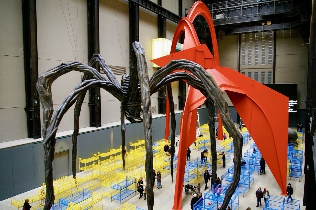 Giant spider-like sculpture in a large industrial hall filled with yellow and blue metal bunk bed frames amongst which people wander