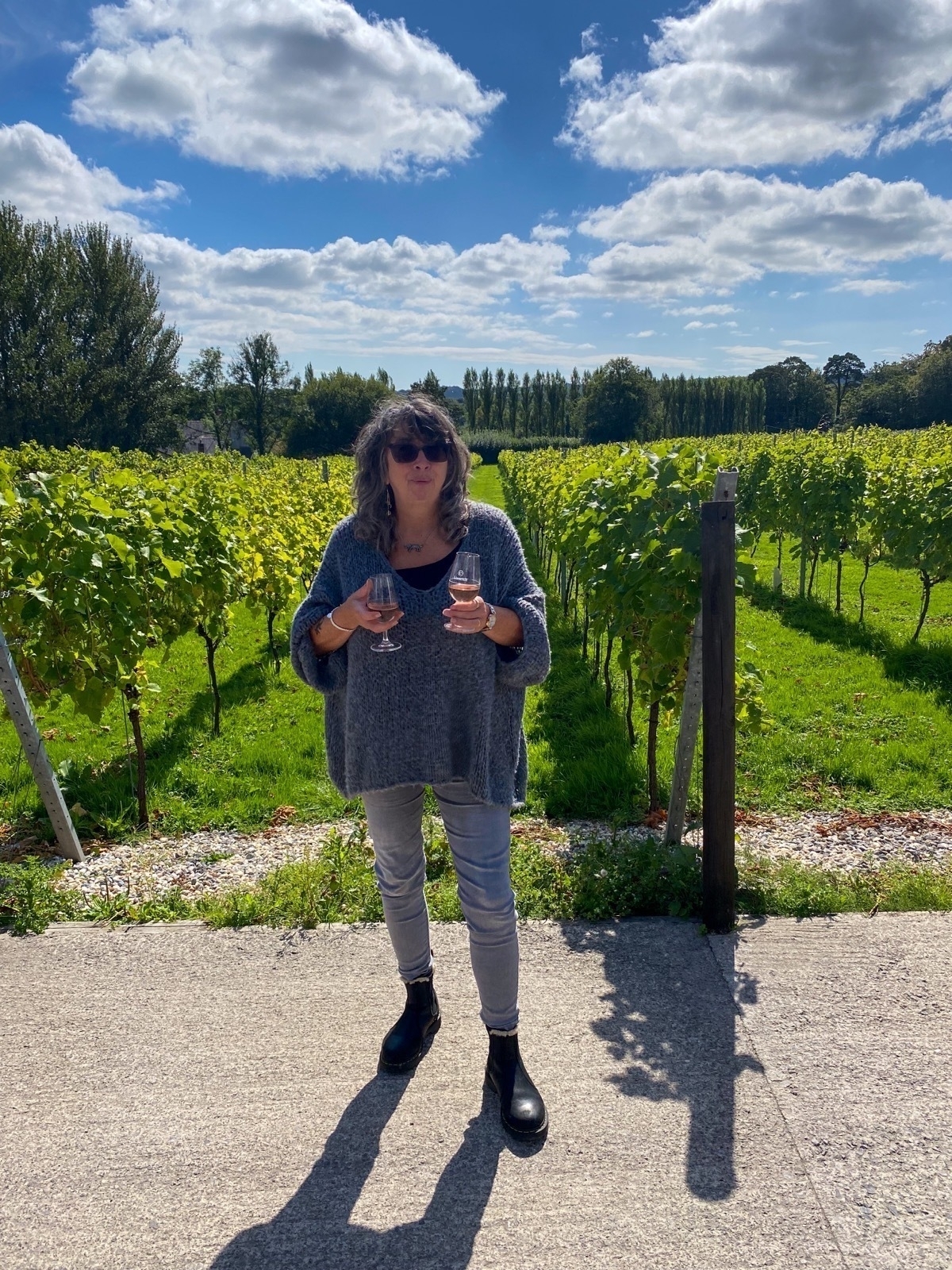 A person is standing in a vineyard holding two glasses with a sunny blue sky and lush green vines in the background.