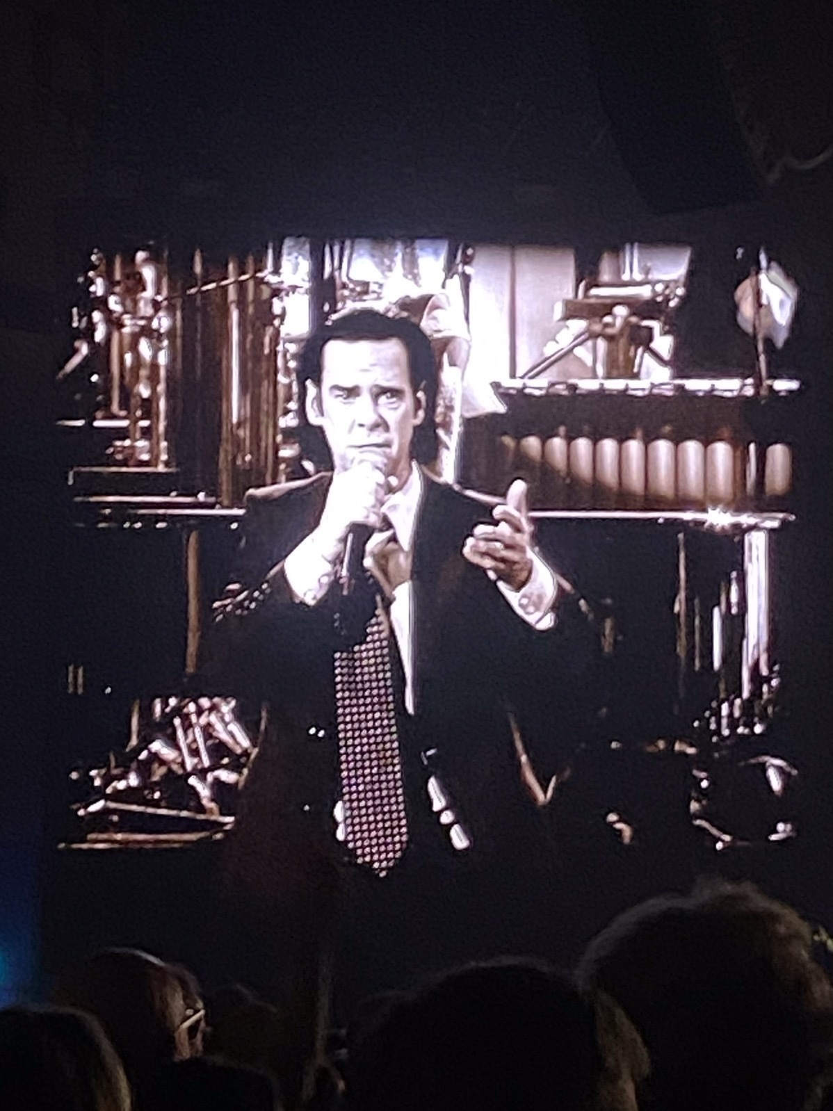 A person in a suit and tie is singing into a microphone on stage, with musical instruments in the background. The image appears to be a large screen projection at a concert with a crowd visible at the bottom. The colors are in a sepia tone