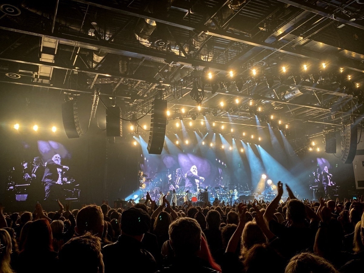 A large concert venue filled with a crowd. The stage is illuminated with bright lights and features musicians performing. Large screens display the performers, with one showing a person holding a microphone. Concertgoers are silhouetted in the foreground, some with raised hands