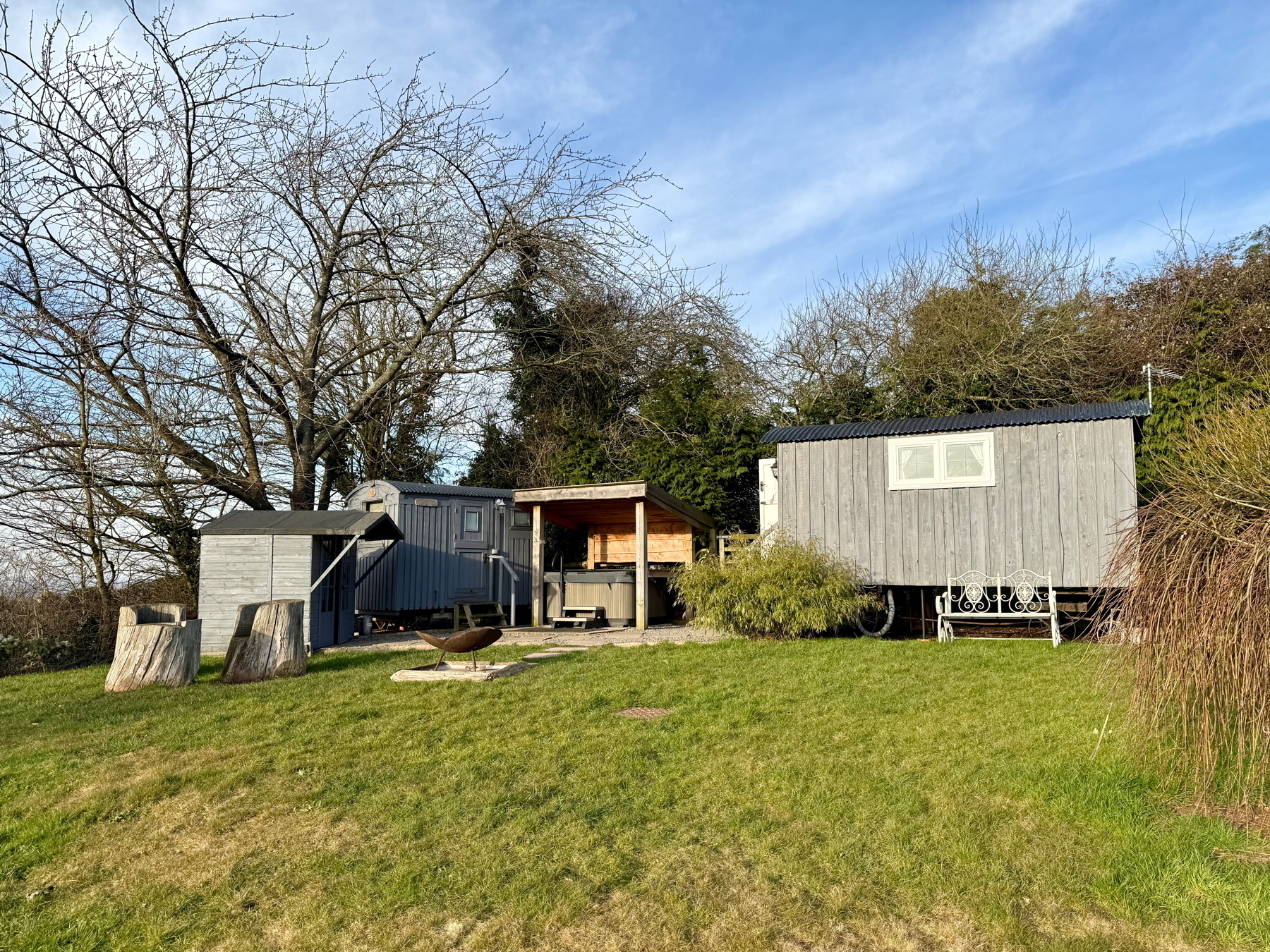 Auto-generated description: A small, rustic cabin with an adjacent shed and outdoor seating area is situated on a grassy hillside surrounded by trees.