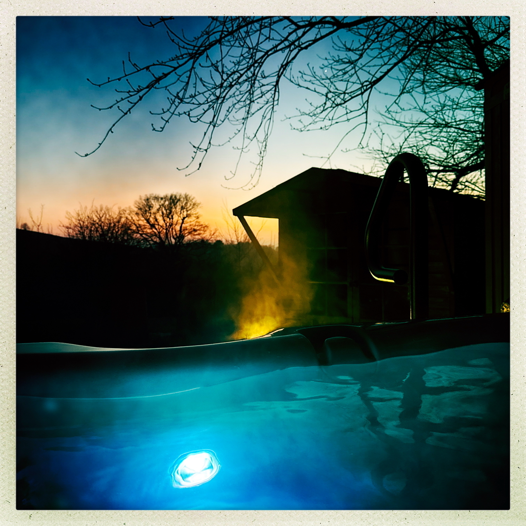 Auto-generated description: A hot tub with a glowing light sits outdoors at sunset, surrounded by trees with silhouetted branches.