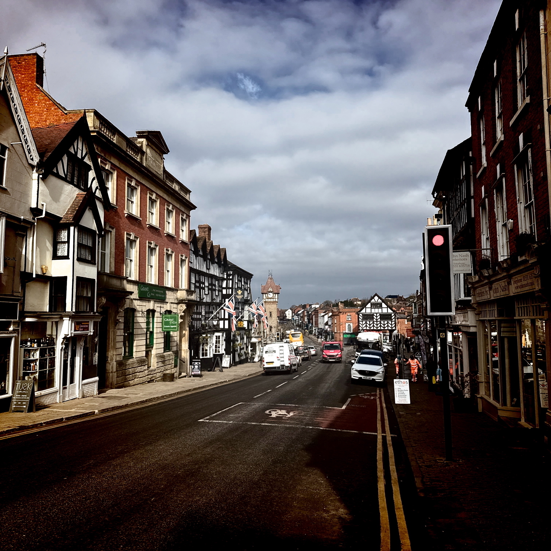 Auto-generated description: A busy town street features historic buildings, a few pedestrians, parked cars, and a cloudy sky overhead.