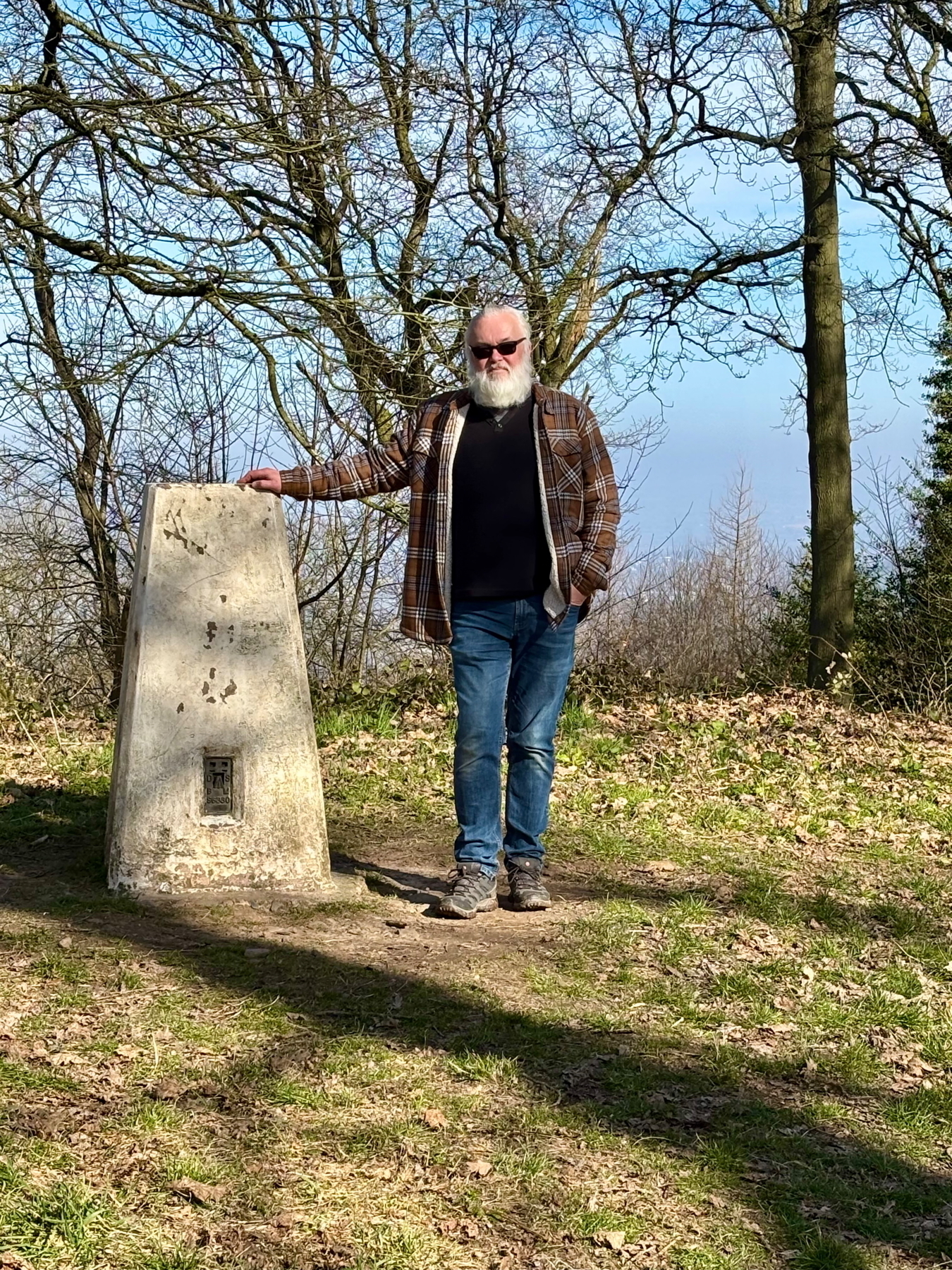 Auto-generated description: A man with a beard is standing next to a stone marker on a hill with leafless trees around.