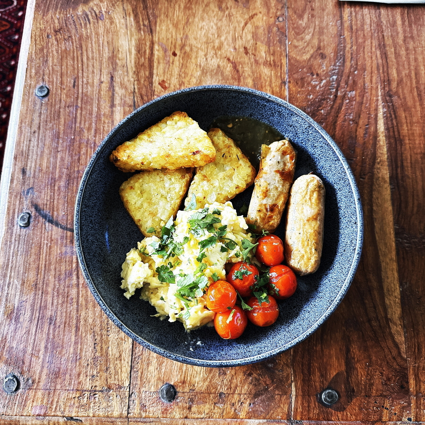 Auto-generated description: A bowl on a wooden table contains hash browns, a sausage, scrambled eggs, and cherry tomatoes.