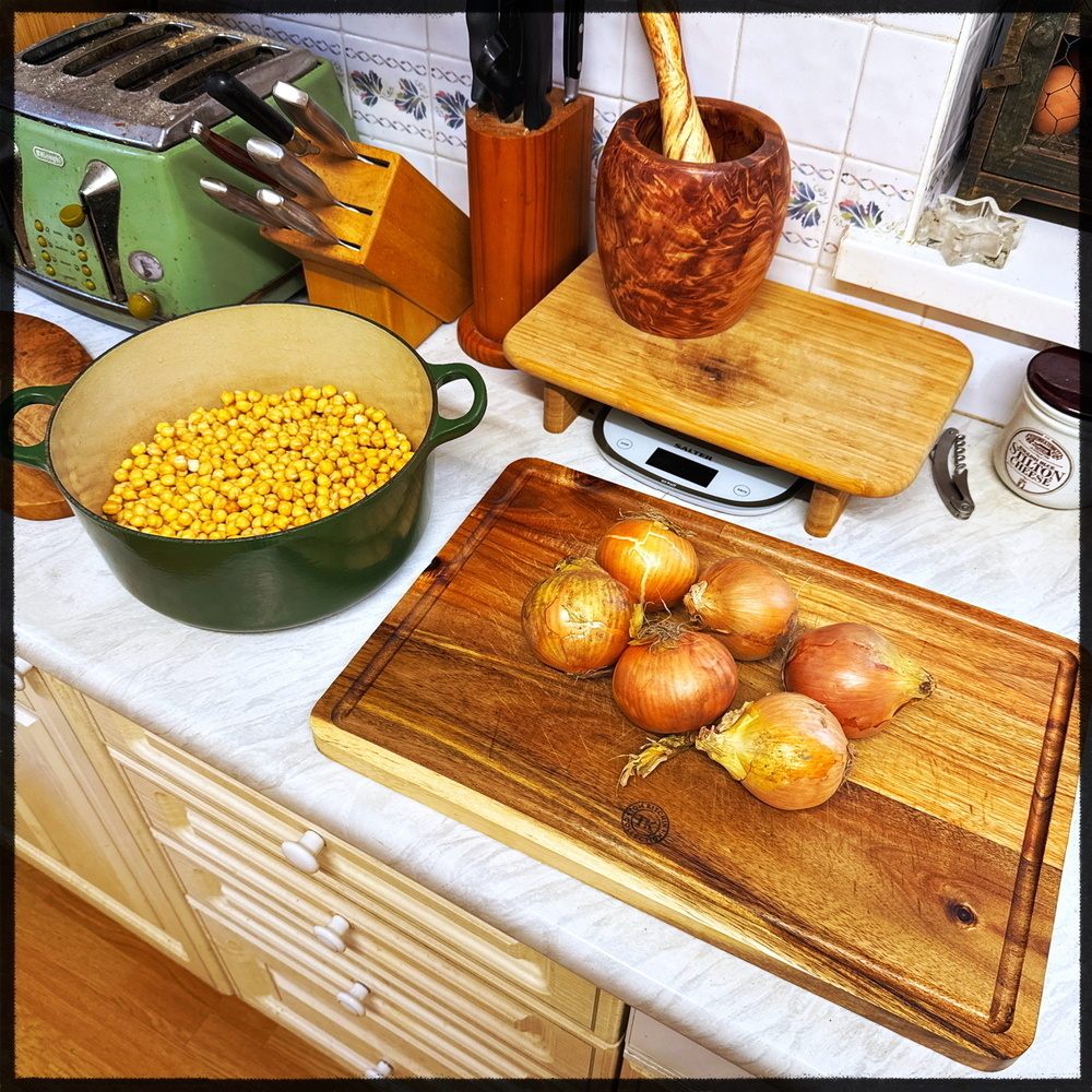 A kitchen countertop displays a wooden cutting board with onions, a green pot filled with chickpeas, a wooden mortar and pestle, knives in a block, and a toaster.