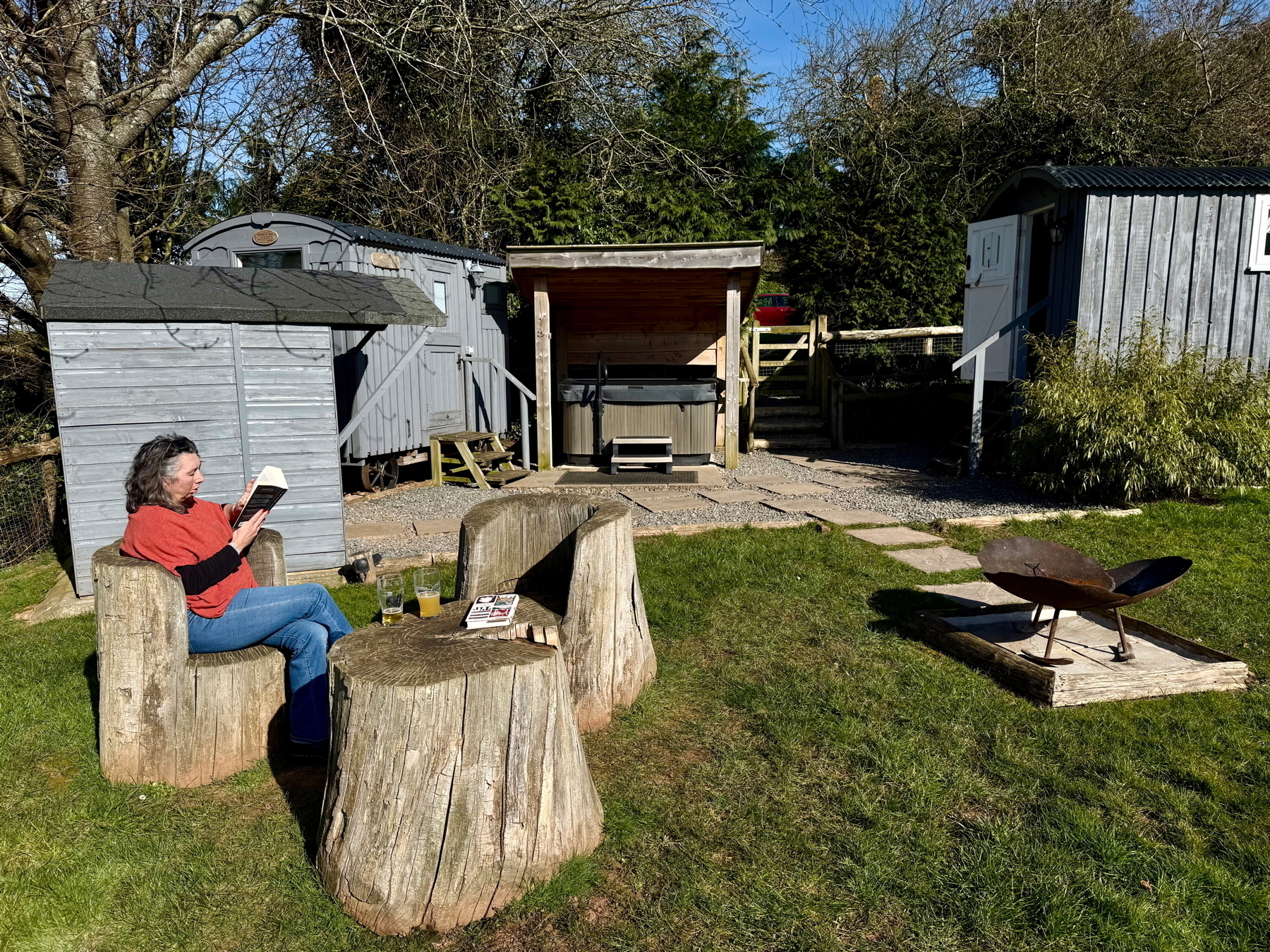 Auto-generated description: A person is sitting on a carved wooden stump in a garden, reading a book, surrounded by rustic wooden cabins and greenery.