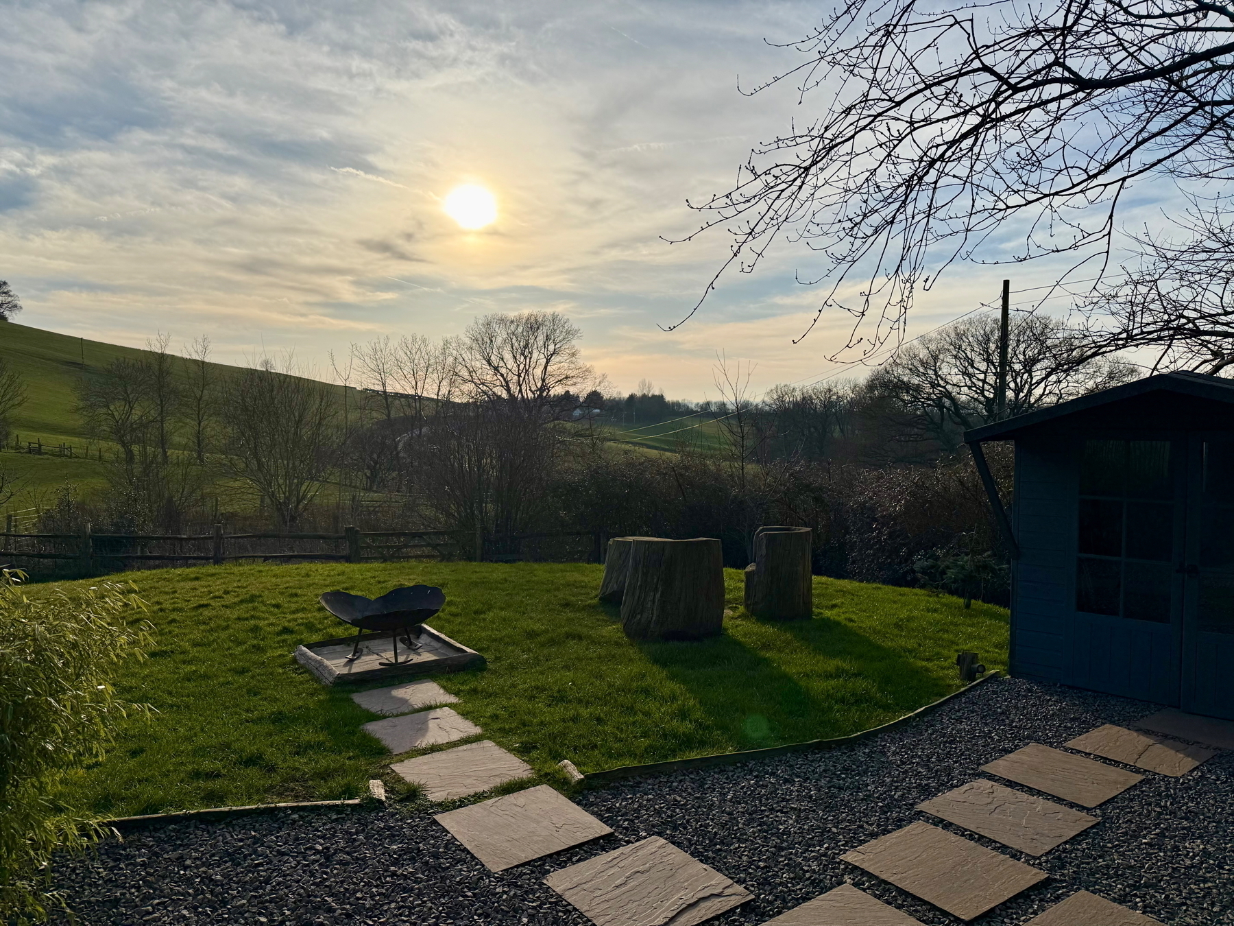 Auto-generated description: A serene garden scene features a grassy area with large tree stumps, a fire pit, and a stone path under a sunlit sky.