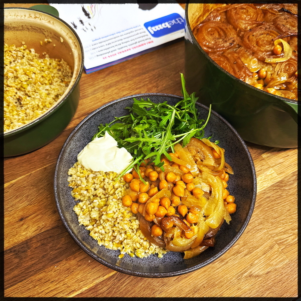 A bowl of grains topped with chickpeas, caramelized onions, arugula, and a dollop of sour cream is placed next to pots containing cooked grains and a dish with more caramelized onions.