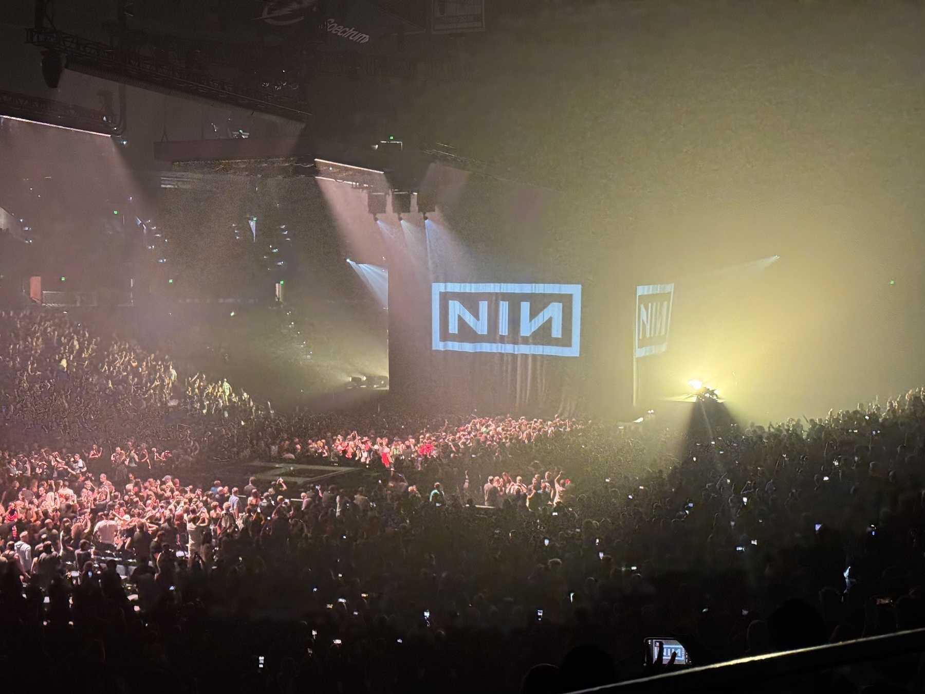 A large crowd is gathered at an indoor concert with a stage displaying the "NIN" logo under dramatic lighting.