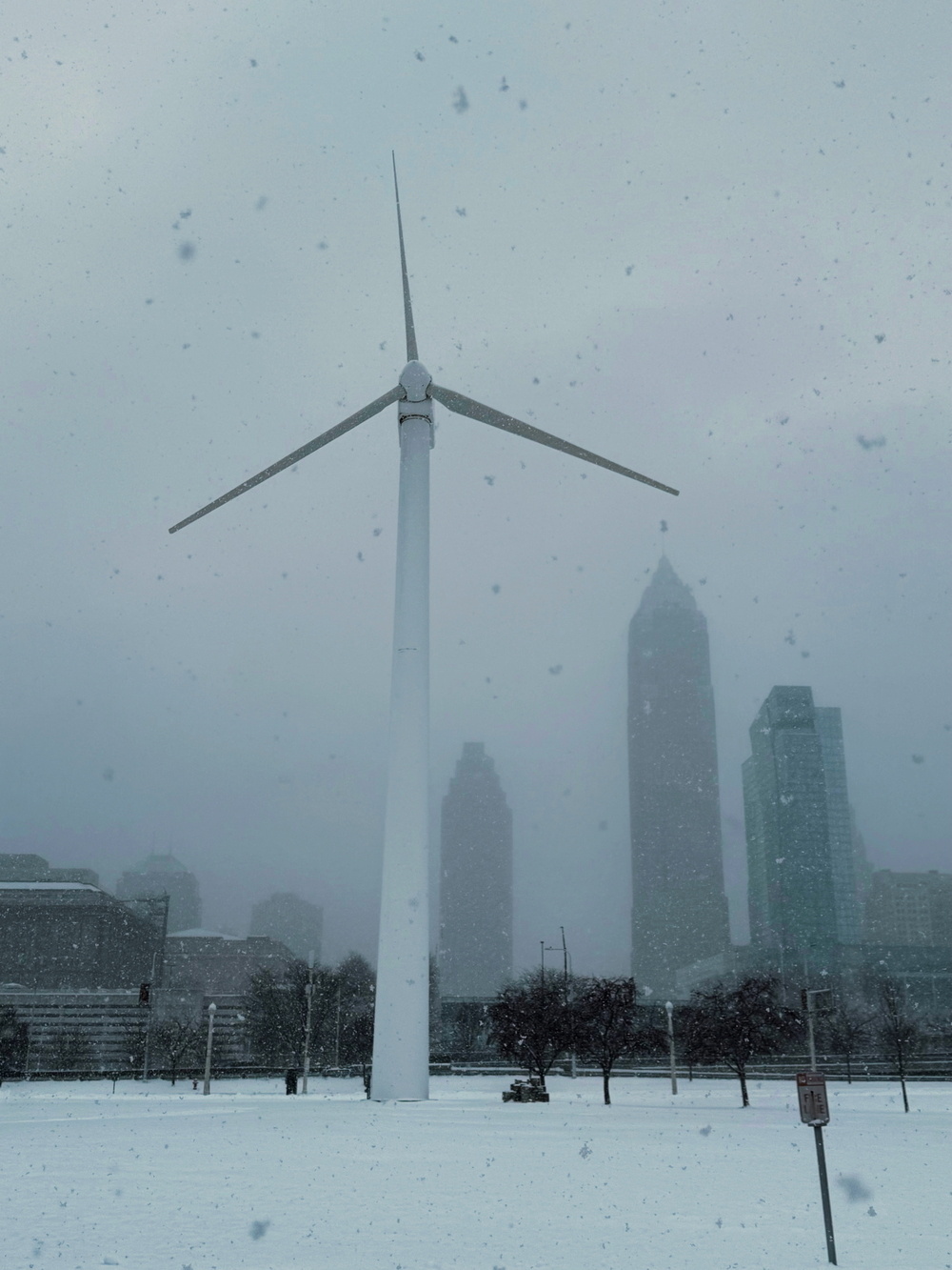 A wind turbine stands tall amidst a snowy landscape with skyscrapers visible in the foggy background.