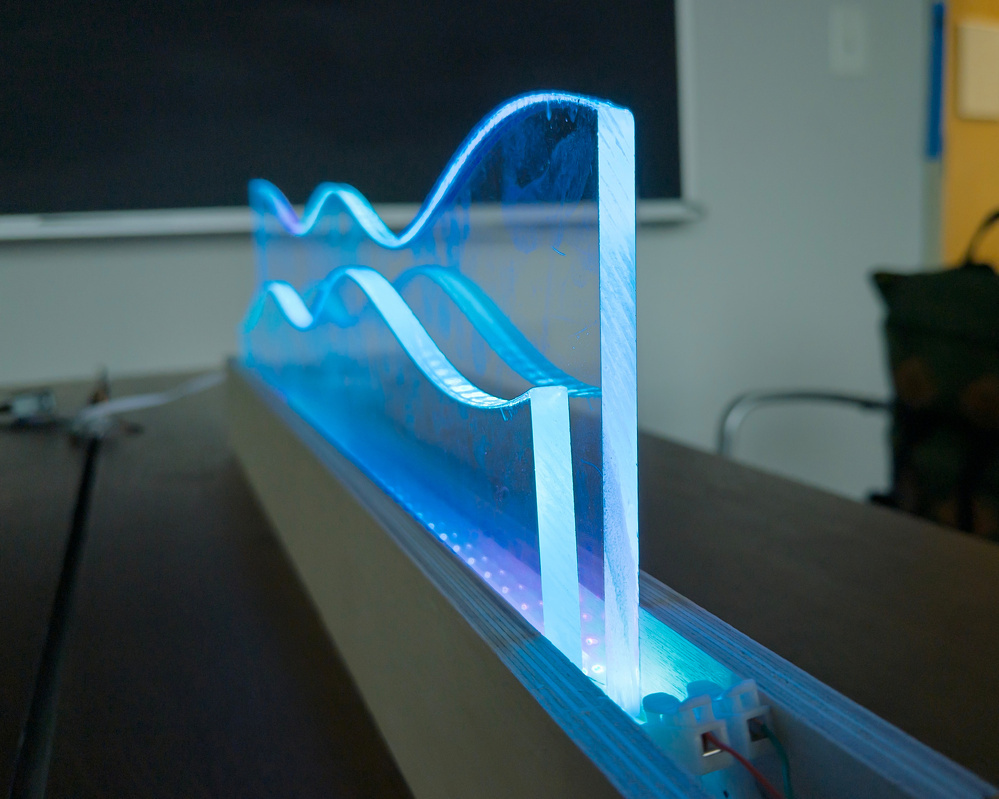 A glowing, wavy acrylic sculpture is mounted on a base, emitting a blue light.