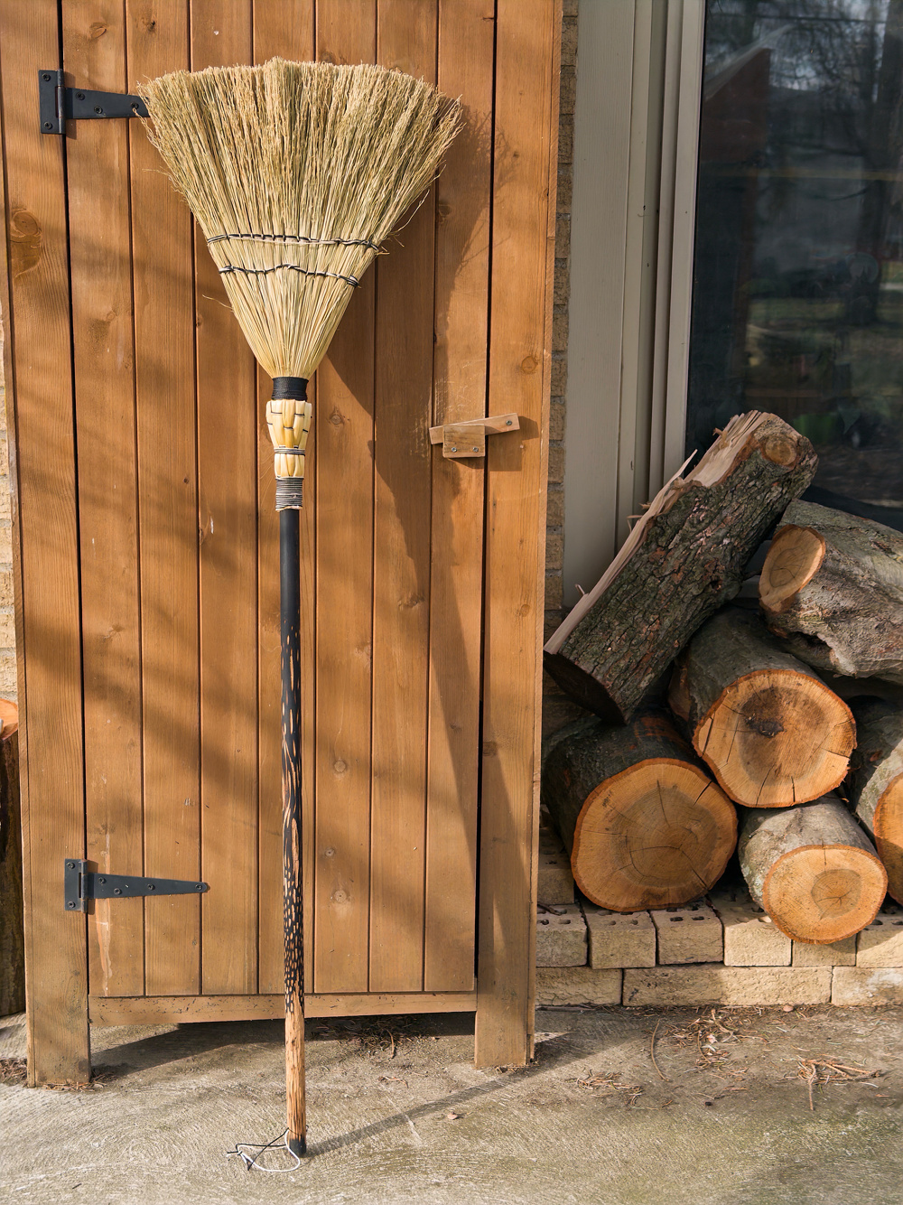 A large broom with a wooden handle is resting against a wooden door, with stacked firewood nearby.