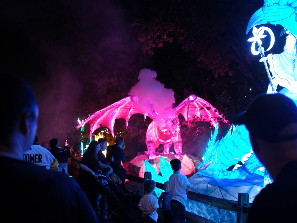 A vibrant dragon sculpture glows in various colors amidst smoke, surrounded by people observing the outdoor scene.