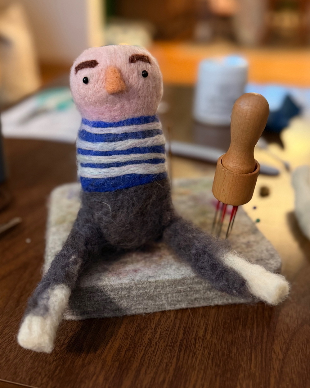 A handmade felted figure with a striped shirt and seated posture is positioned on a table with felting tools nearby.