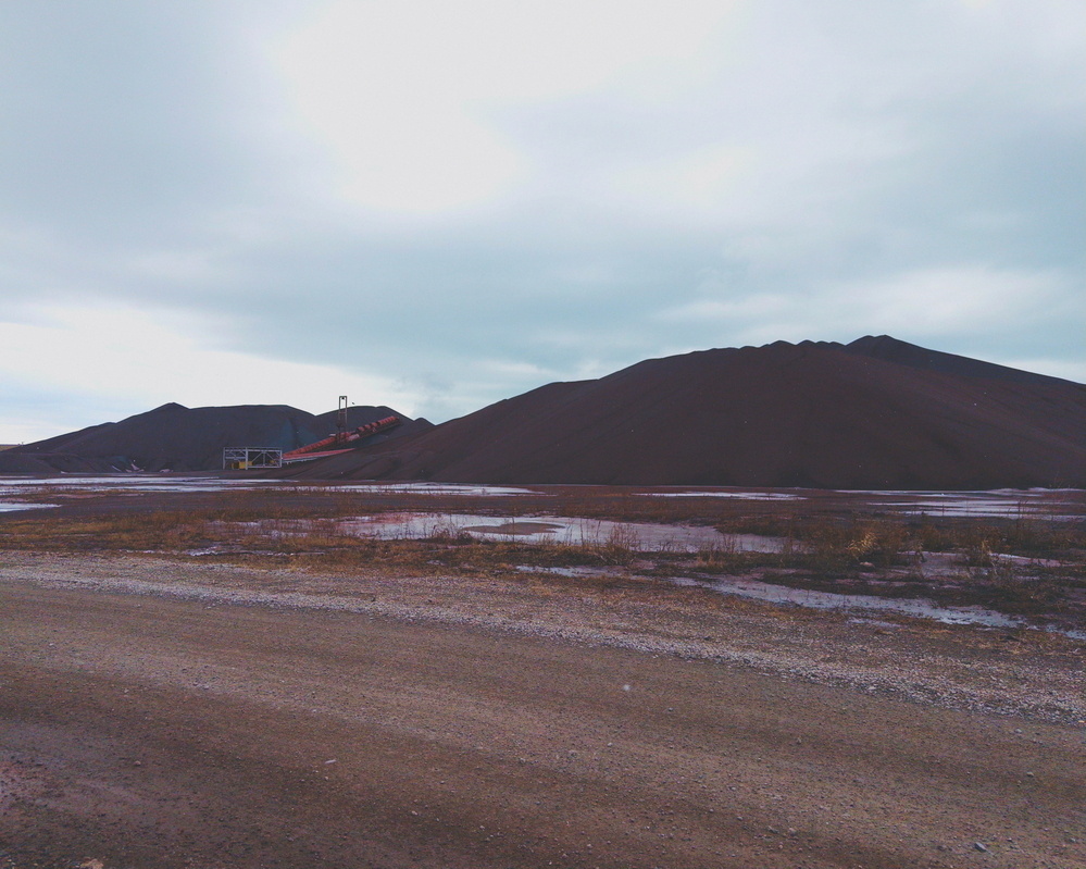 A barren landscape features large piles of dark soil or gravel with an overcast sky above.