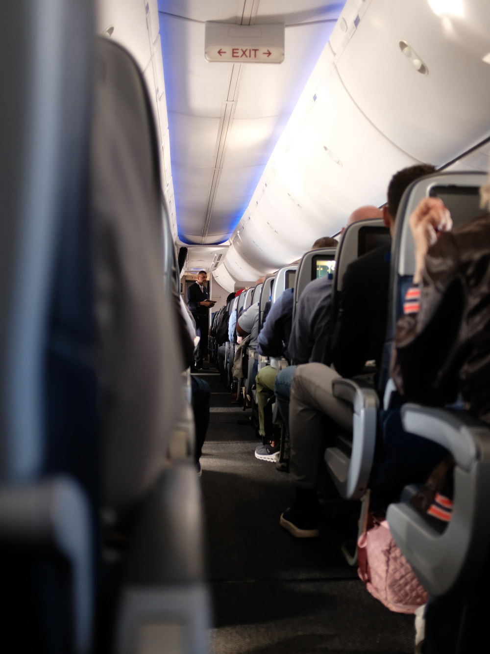 A view down the aisle of an airplane cabin shows passengers seated and an EXIT sign overhead.