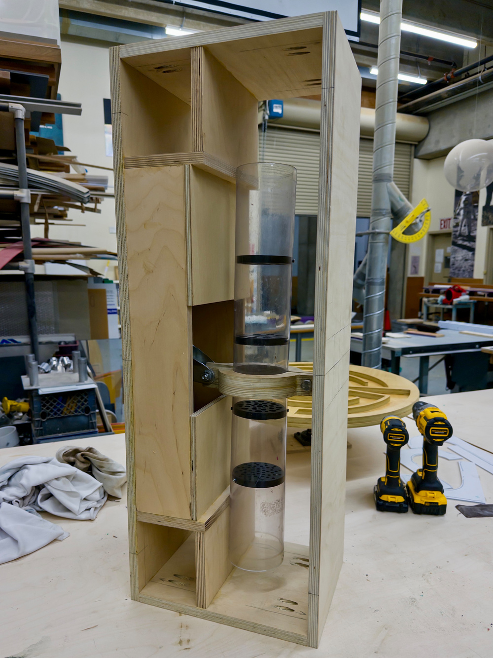 A wooden structure contains vertically stacked clear tubes and is surrounded by tools and workshop equipment.