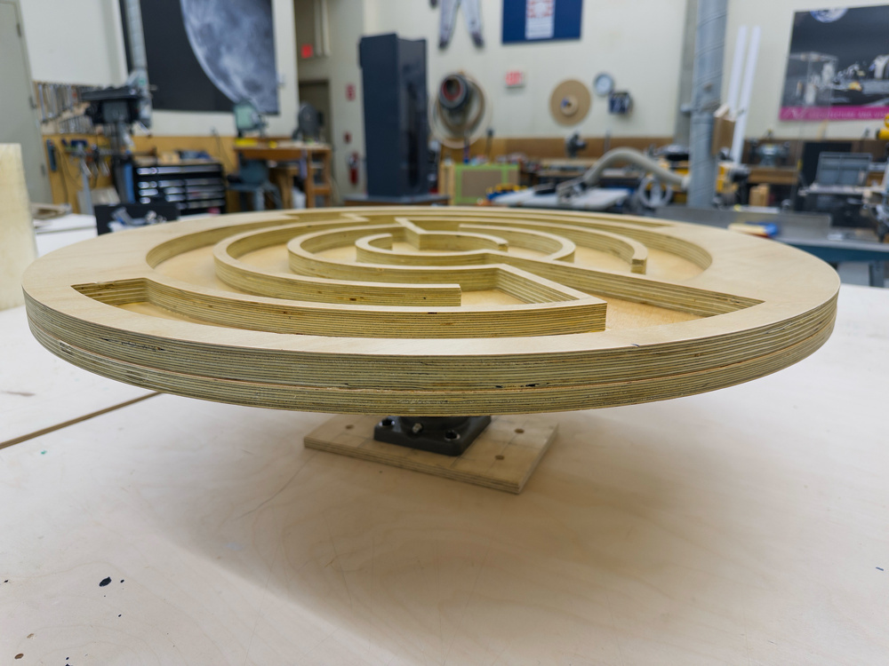 A wooden, circular maze is mounted on a rotating platform in a workshop setting.