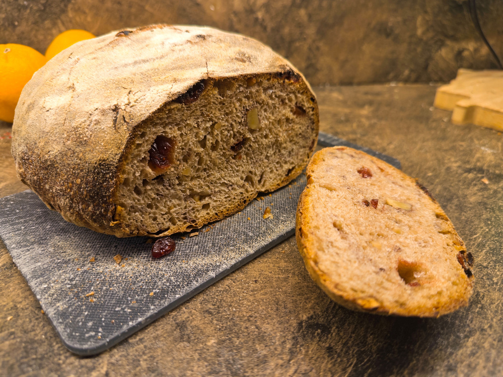 A loaf of rustic bread with cranberries and nuts is partially sliced on a cutting board.