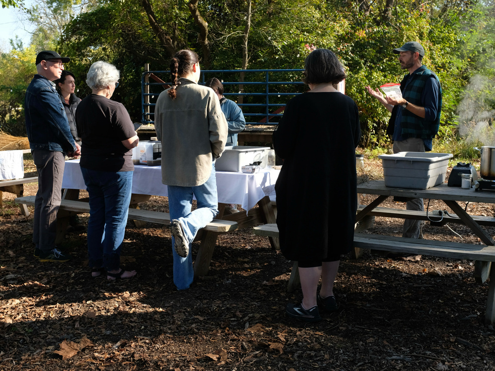A group of people stand around picnic tables outdoors, where a man is holding a booklet or pamphlet.