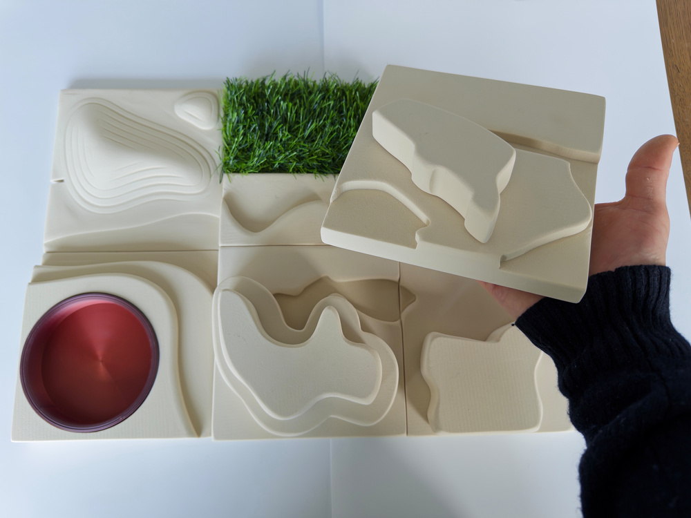 A person is holding a puzzle piece above a set of textured tiles, including one with artificial grass and another with a circular red feature.