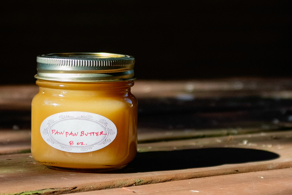 A jar of pawpaw butter labeled 8 oz. sits on a wooden surface, partially in shadow.