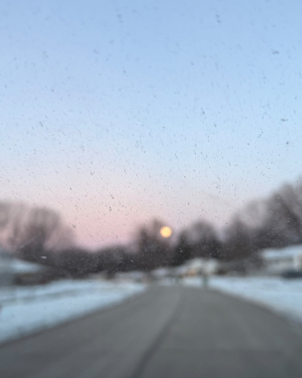 A blurred view of a snowy road at dusk is seen through a speckled window, with trees and a faint moon on the horizon.