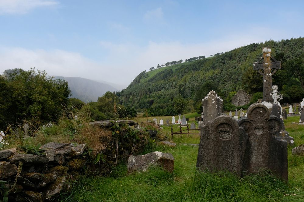 A graveyard with weathered tombstones is set against a backdrop of lush green hills and a cloudy sky.