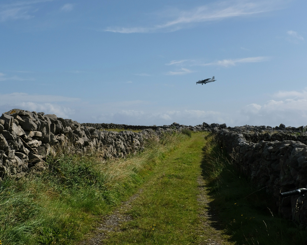 A small plane flies over a narrow grassy path lined with stone walls under a clear blue sky.