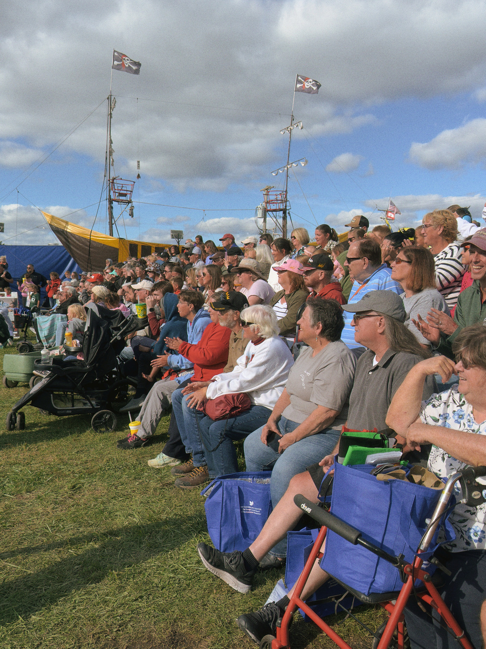 A crowd of people sits on bleachers watching an outdoor event with flags and a ship structure in the background.
