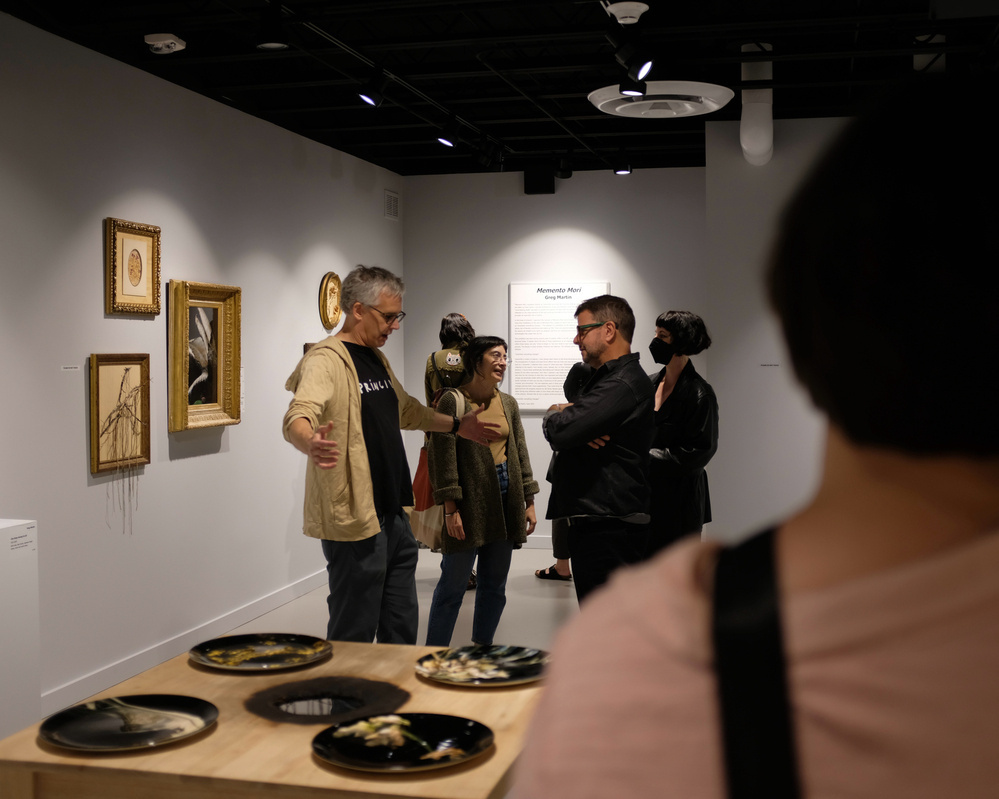 A group of people are gathered in an art gallery, observing framed artwork on the walls and ceramics on a table.
