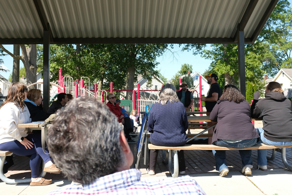 A group of people are sitting on benches under a pavilion, listening to someone speaking in front of a playground.