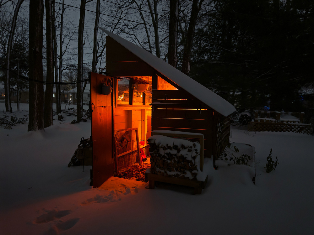 A snow-covered backyard features a small wooden shed emitting a warm, orange glow from inside, contrasting with the dark, snowy surroundings.