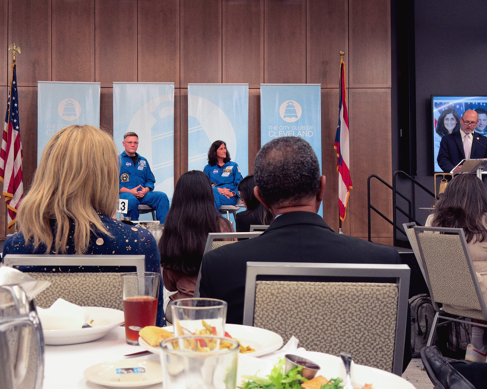 Two individuals in blue uniforms are seated on a stage at an event, with an audience and a speaker at a podium nearby.