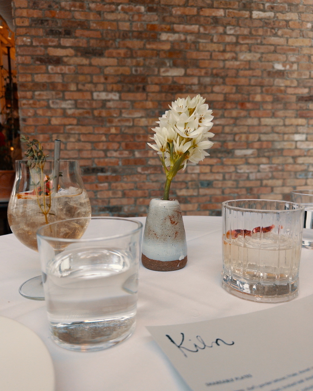 A table set with drinks, a small vase of white flowers, and a menu is placed against a brick wall backdrop.