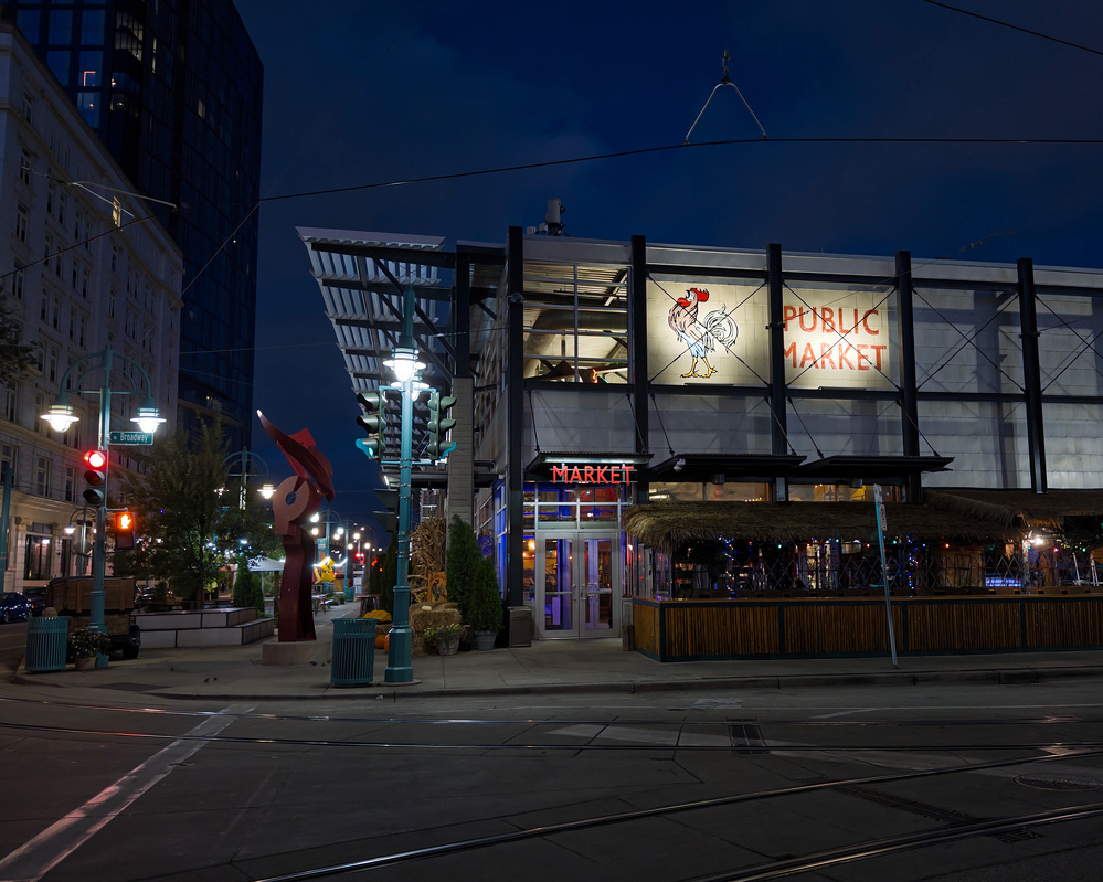 A lit-up public market building and street scene are visible at night, featuring a tall sculpture and surrounding urban architecture.