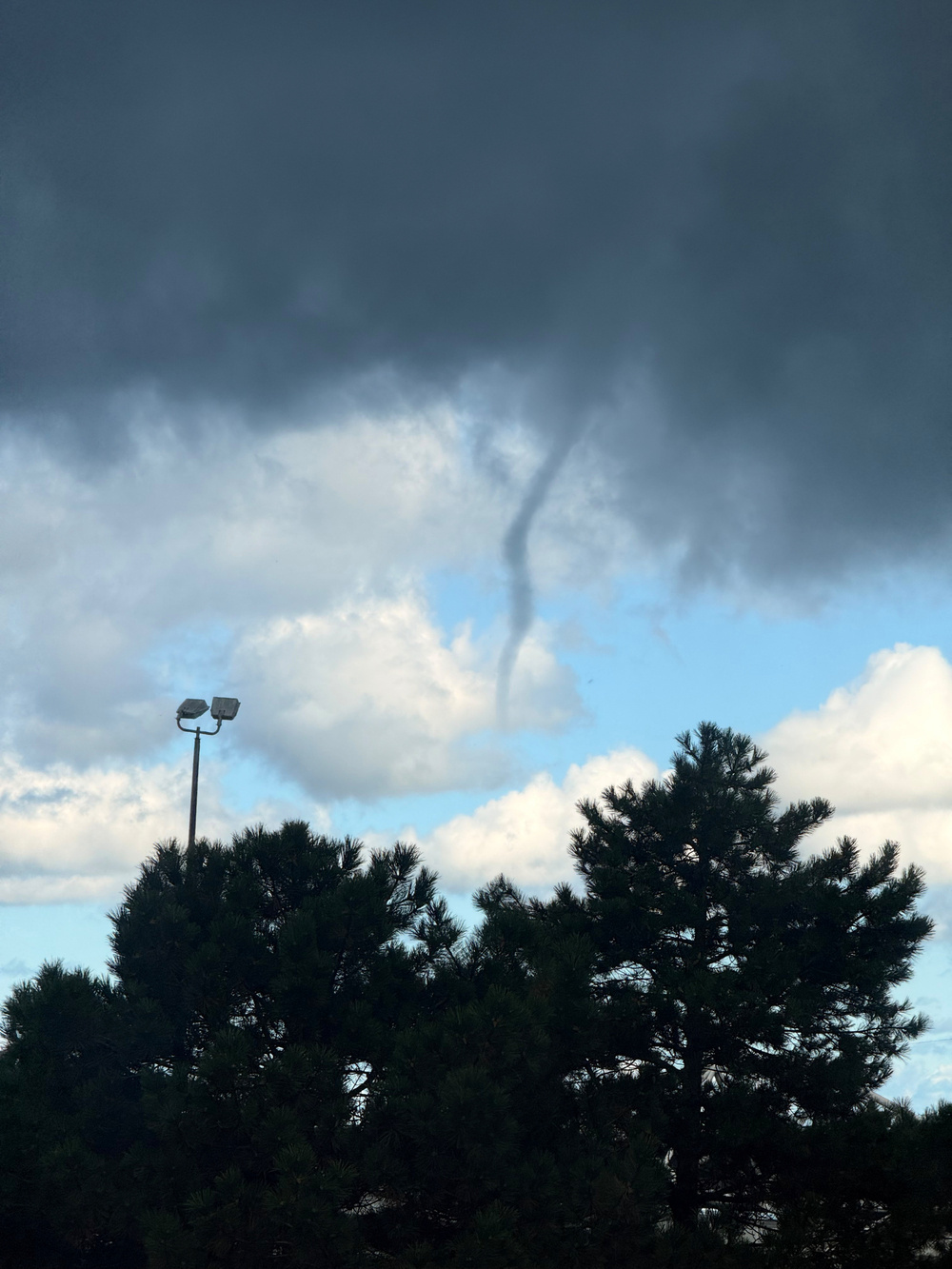 A funnel cloud extends from dark, ominous clouds above a landscape with trees and a light pole.