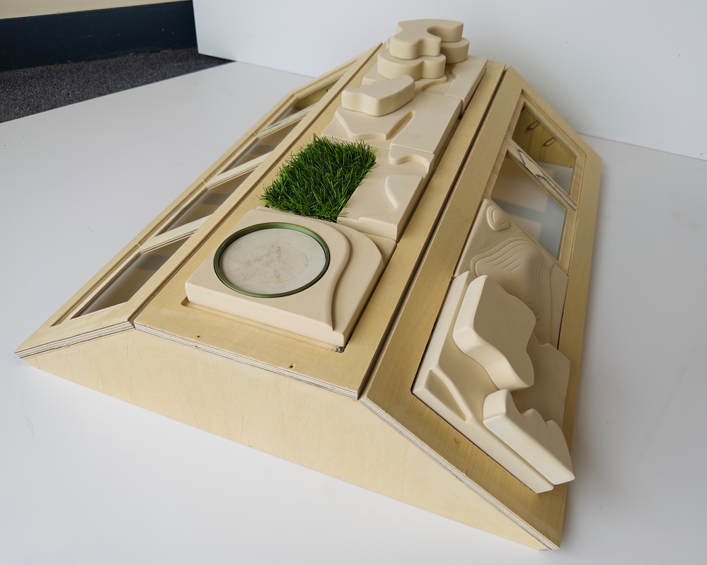 A triangular architectural model features a variety of natural elements and terrain textures, including artificial grass and water pathways, on a sloped wooden base.
